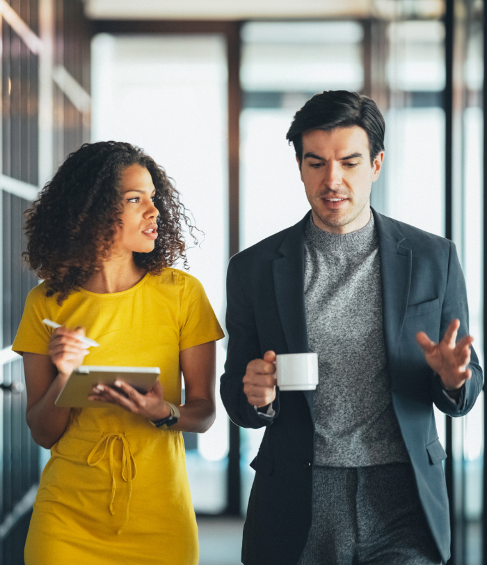 Male and female colleague walking down office hallway holding coffee and digital tablet respectively